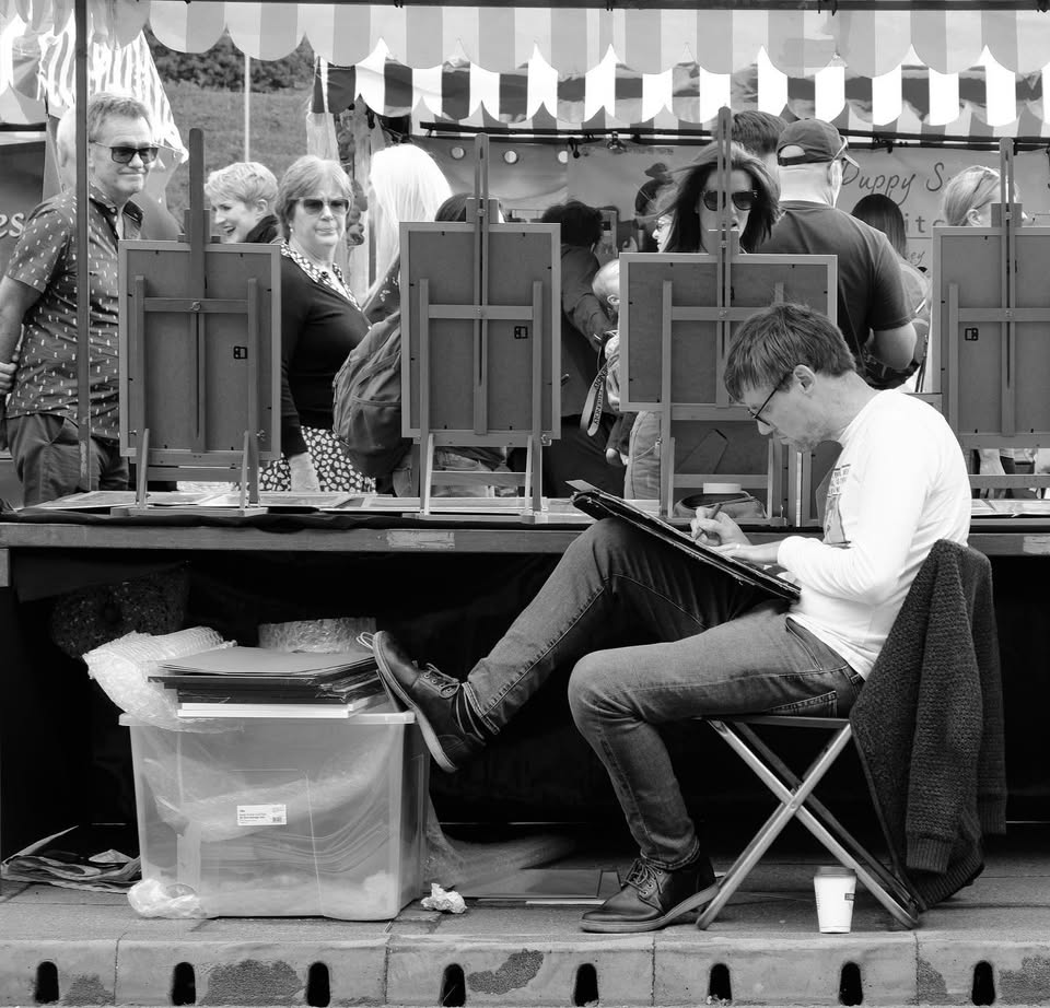 An image of Ben Holland at one of his market stalls. It is in black and white and he is leaning in a chair, drawing an image we can't see while members of the public look at his work.