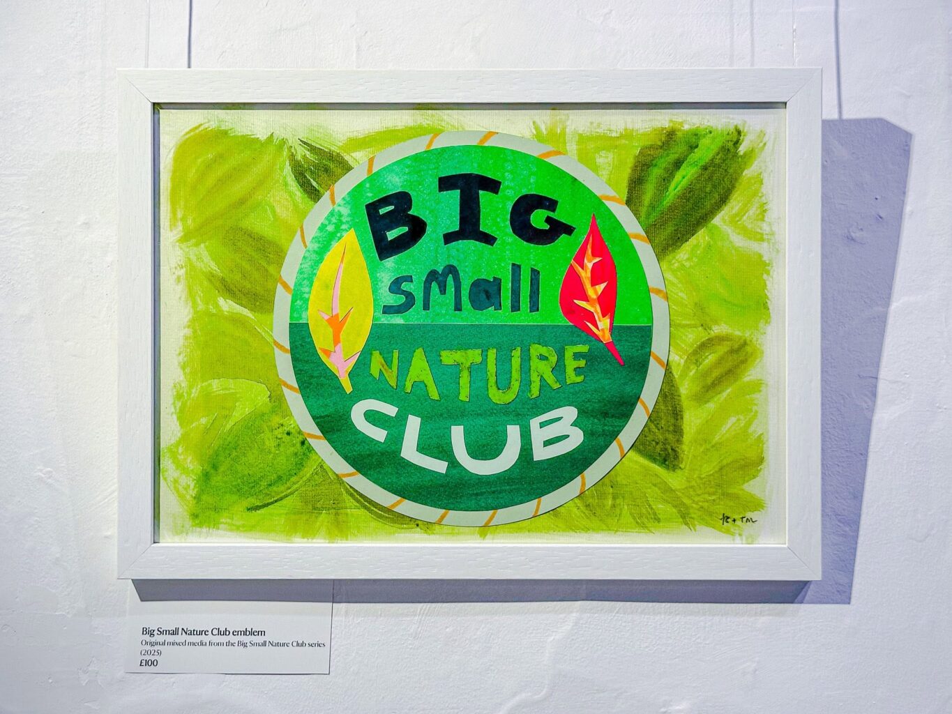 A framed artwork on a white painted wall. A sign underneath it reads 'Big Small Nature Club Emblem. Original mixed media from the Big Small Nature Club series (2025). £100.'. The artwork is a circular emblem made from collaged paper, with 'Big Small Nature Club' in big blocky test accompanied by two bright paper leaves. The background is a messy mix of greens that emulate a carpet of leaves.