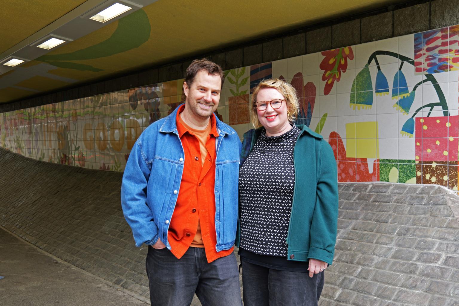 Two people stand next to each-other in an underpass decorated with a fun and lively mural. On the left is Tom Madge, dressed in a bright orange shirt and denim jacket. On the right is Josie Brookes, dressed in a black shirt with white hearts and bottle green jacket.