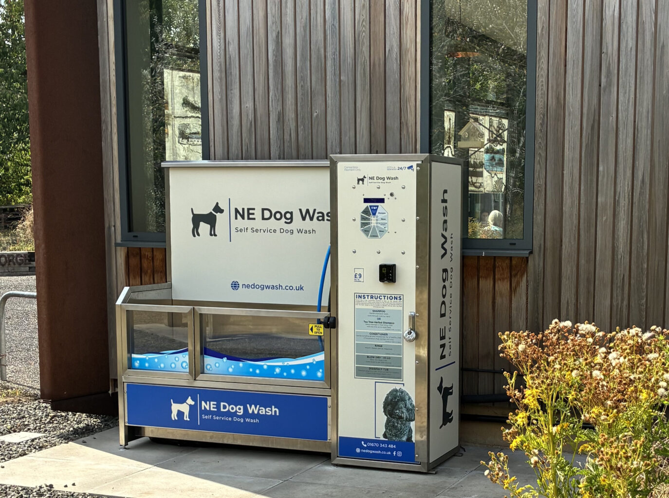 An image of a dog-washing station, which includes and enclosed tub, hose shower and signage explaining how it operates and pricing. The logo of NE Dog Wash, which includes a logo that is the silhouette of a dog, is printed large on it's back wall. In the background is the Land of Oak & Iron Heritage Centre